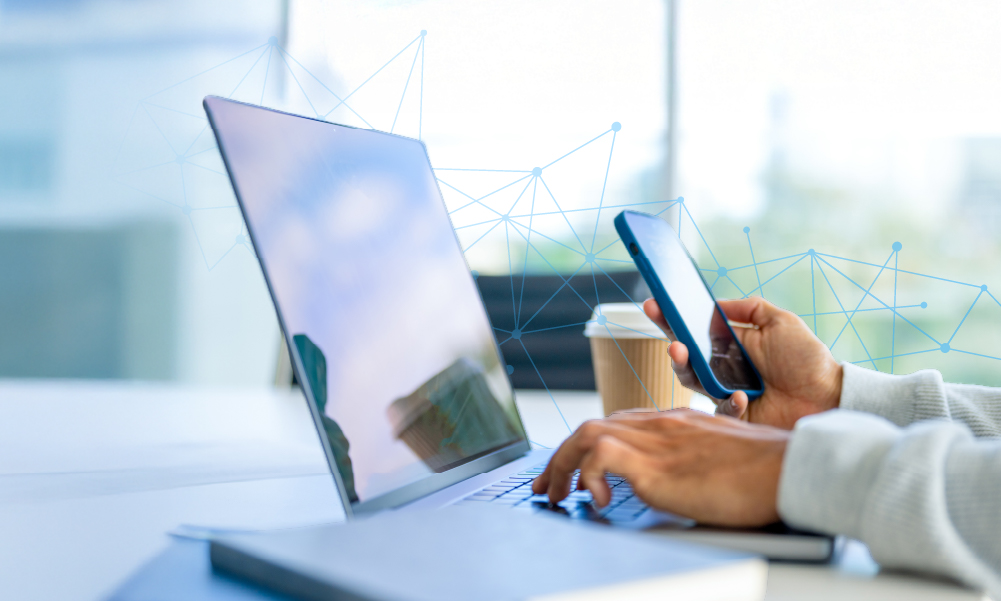 Person using laptop and smartphone at desk with coffee cup and digital network overlay.