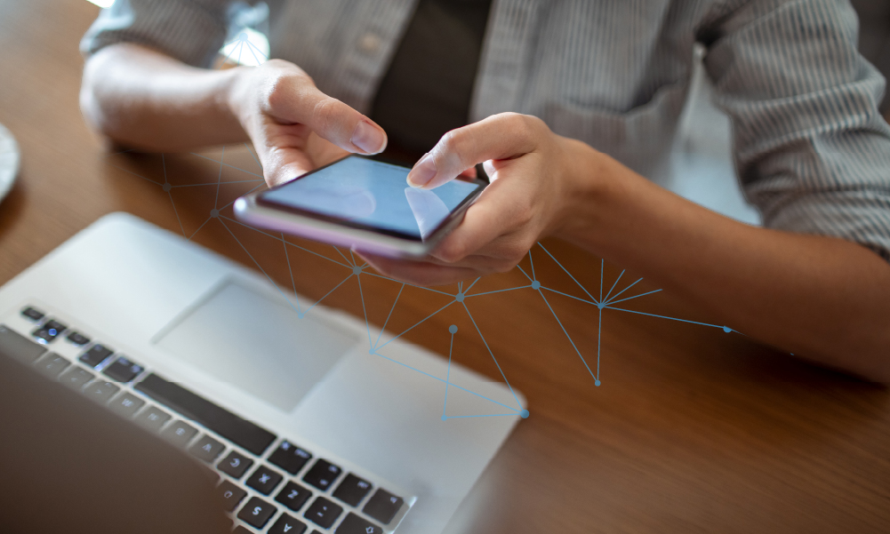 Person using smartphone above laptop on wooden desk with digital network overlay.